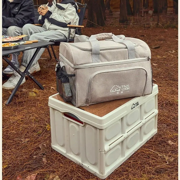 Two gray carrying bags on top of a white storage box in an outdoor setting with people and chairs in the background.