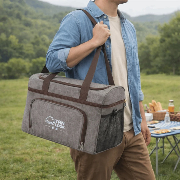 Person carrying a gray and brown cooler bag outdoors with a picnic setup in the background.