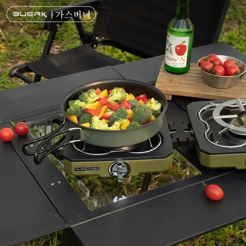 Outdoor cooking setup with a portable stove, pan with vegetables, and a bottle of beer on a grassy area.