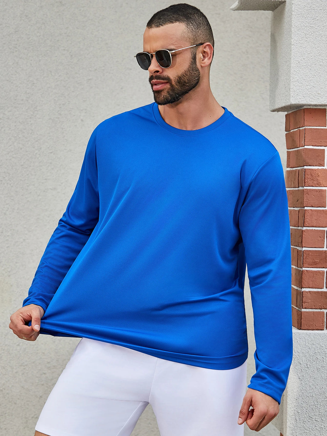 Man wearing a blue long-sleeve shirt and white shorts standing against a light gray wall.