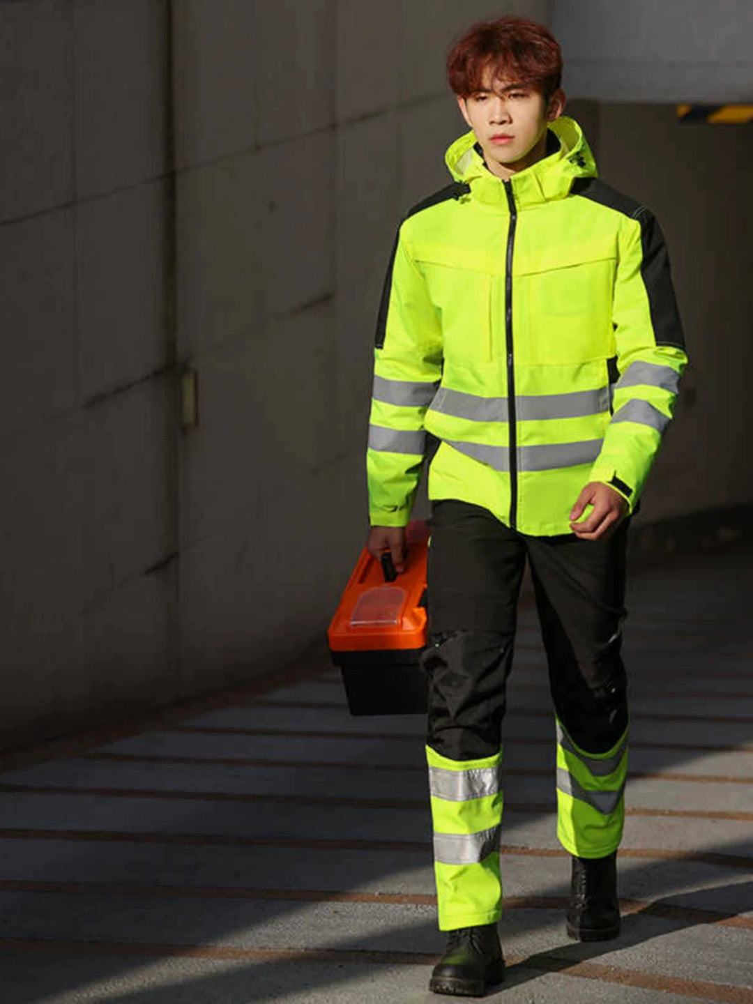 Person wearing a high-visibility safety jacket and pants, holding a toolbox.