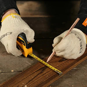 Person measuring a wooden plank with a tape measure and pencil, wearing gloves.