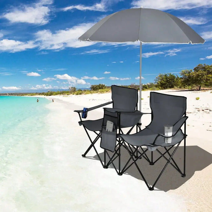 The double chair set sits on a sunny beach with the umbrella casting shade over the seats, offering portable comfort with cupholders and a center cooler between the seats.
