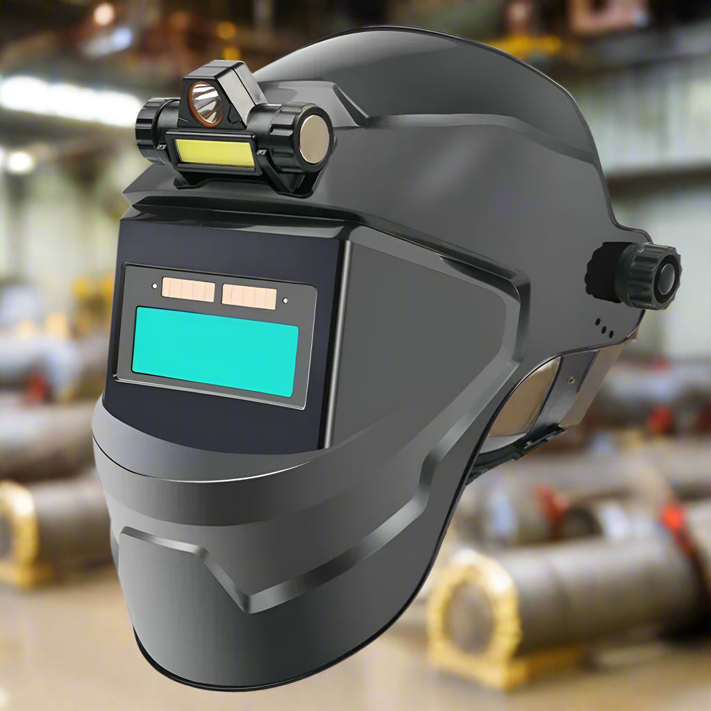 Black auto-darkening welding helmet with a classic lens and built-in LED light, displayed in an industrial workshop with welding equipment in the background.