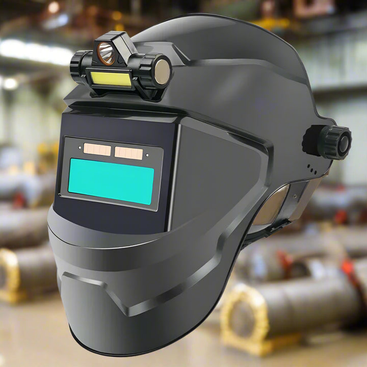 Black auto-darkening welding helmet with a classic lens and built-in LED light, displayed in an industrial workshop with welding equipment in the background.