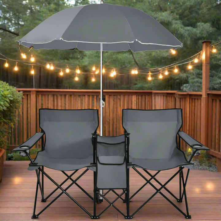 Dual gray folding chairs with a built-in center cooler and matching umbrella are set up on a wooden deck with warm string lights creating a relaxing evening vibe.