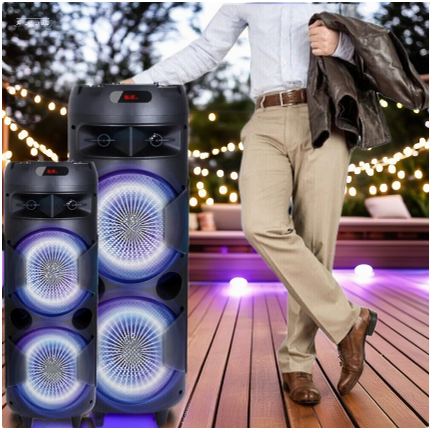 Tall dual speakers with glowing RGB lights set on an outdoor deck under hanging string lights, with a man casually standing nearby holding a jacket.