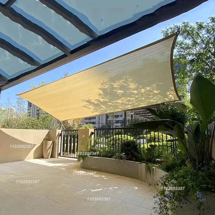 A beige sun shade sail installed above a modern patio, providing cooling shade over concrete and plants, attached with tension ropes under a pergola roof.