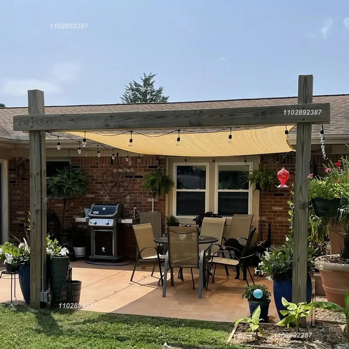 A shade sail mounted over a backyard patio dining area, attached to a pergola. Warm string lights hang underneath, creating a cozy outdoor dining space.