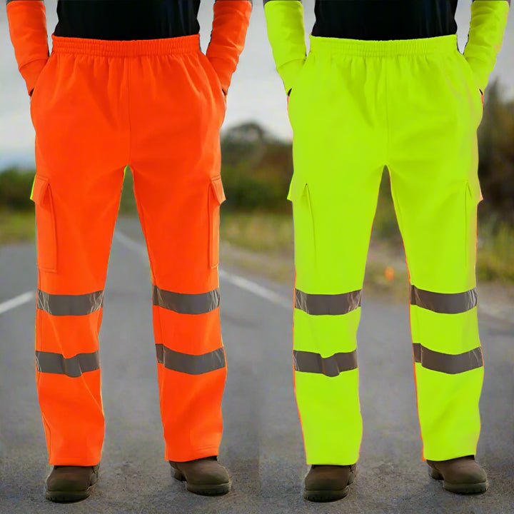 Two workers wearing high-visibility orange and lime safety pants standing outdoors during daylight, with shrubs and foliage in the background.