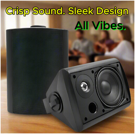 Black stereo speakers on a wooden table with a blurred background of people playing music. Overlaid text reads: "Crisp Sound. Sleek Design. All Vibes."